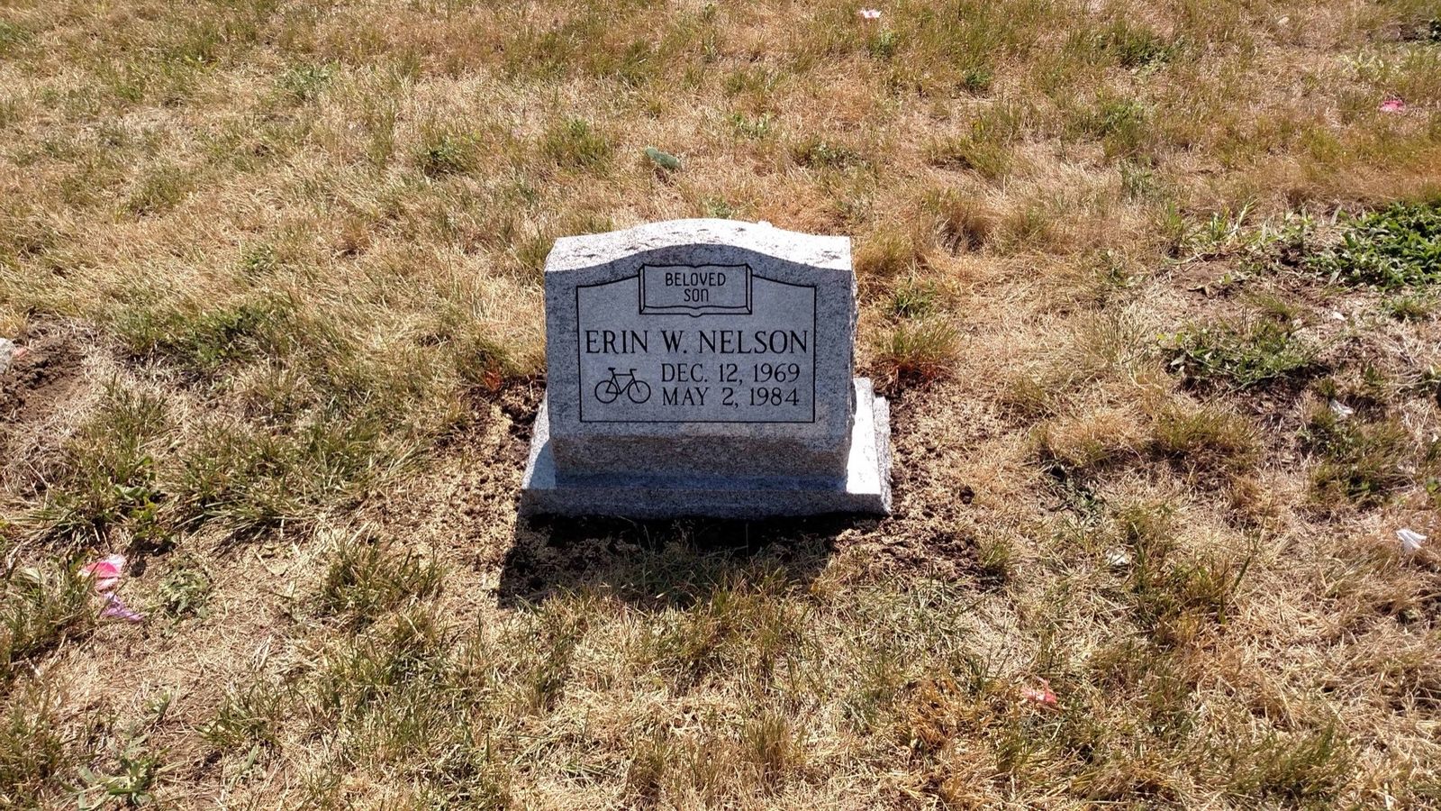 Grey granite upright with a carved bicycle and the words Beloved Son, standing in cemetery grass