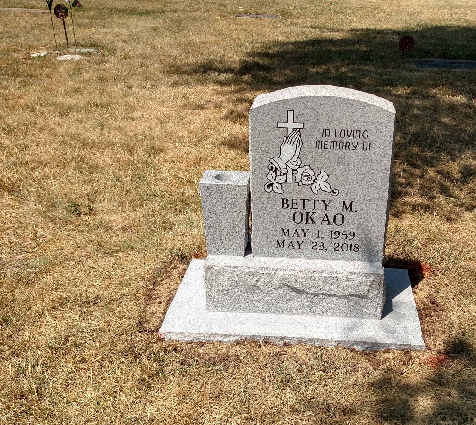 Grey granite upright monument carved with praying hands and a cross, with a granite vase alongside