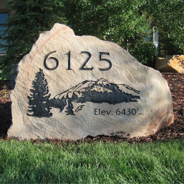 Natural stone boulder carved with a house number and a mountain and pine scene